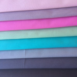 May include: A stack of seven folded fabric swatches arranged in horizontal layers. The colours from top to bottom are light pink, lavender, bright pink, mint green, turquoise, slate blue, and dark charcoal grey, all featuring a visible woven texture.