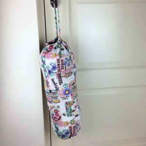 May include: A cylindrical fabric bag with a drawstring closure, hanging from a metal hook. The bag features a floral pattern with pink, blue, and green accents, and text that reads "Flower Seeds".