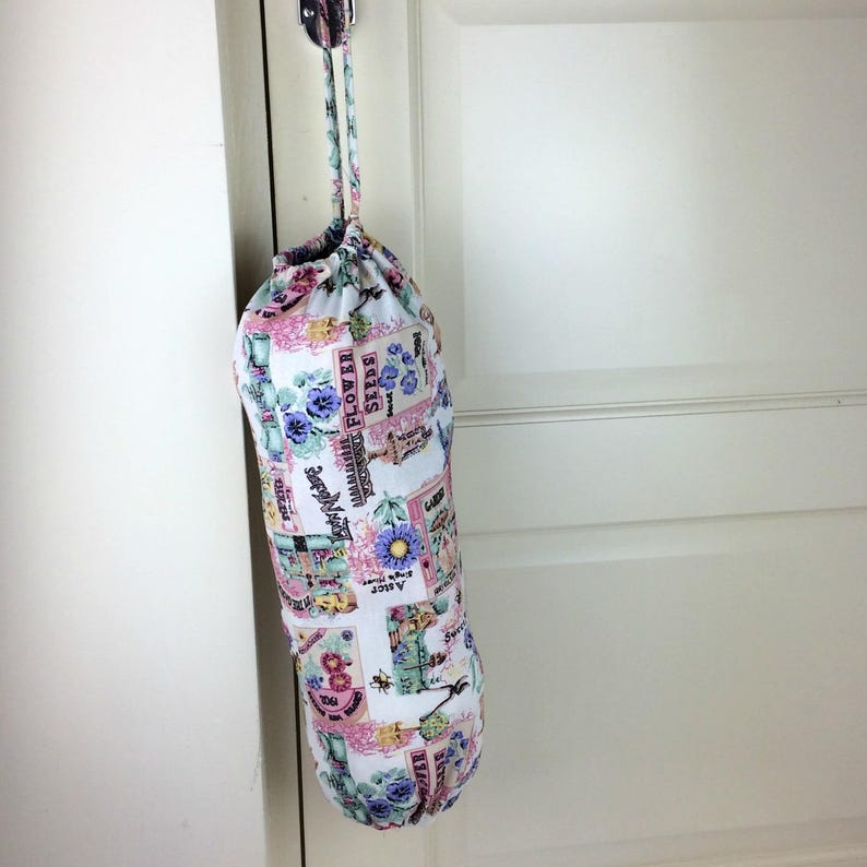 May include: A cylindrical fabric bag with a drawstring closure, hanging from a metal hook. The bag features a floral pattern with pink, blue, and green accents, and text that reads "Flower Seeds".