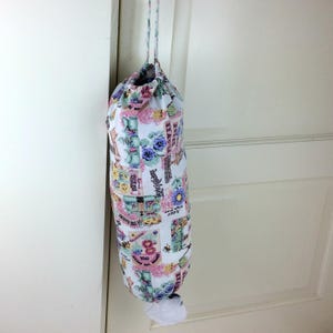May include: A fabric grocery bag dispenser with a drawstring closure, hanging from a door. The bag is white with a floral and vintage postcard print in pink, blue, and green. A white plastic bag is visible at the bottom.