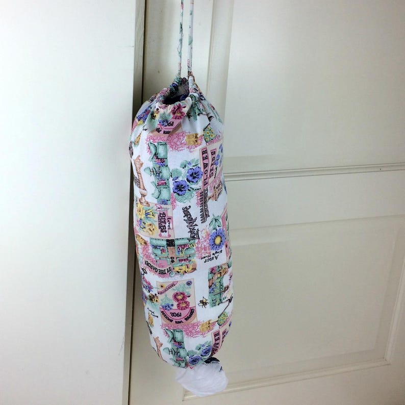 May include: A fabric grocery bag dispenser with a drawstring closure, hanging from a door. The bag is white with a floral and vintage postcard print in pink, blue, and green. A white plastic bag is visible at the bottom.