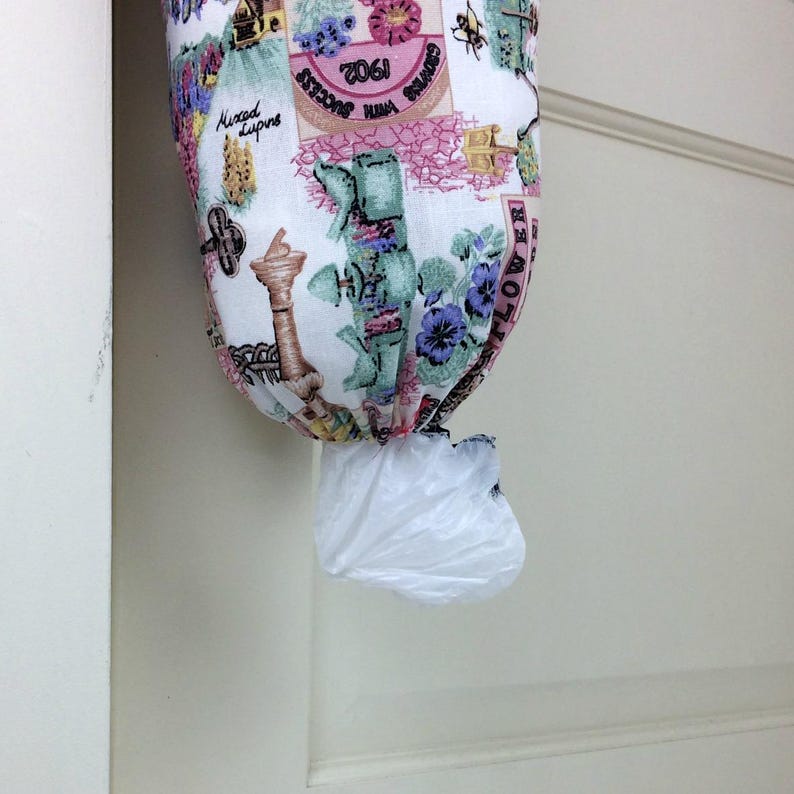 May include: A fabric bag dispenser with a white background. The bag is made of a patterned fabric with floral and architectural designs in shades of pink, green, and blue. A white plastic bag is visible at the bottom.
