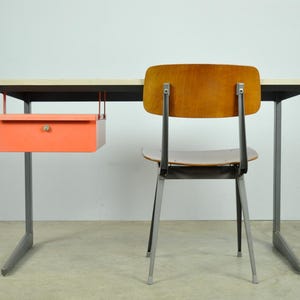 Könnte beinhalten: Ein Schreibtisch im Mid-Century-Stil mit einer hellen Holzplatte, schwarzen Zierleisten und grauen Metallbeinen. Eine orangefarbene Schublade hängt darunter. Ein Holzstuhl mit grauen Metallbeinen steht hinter dem Schreibtisch.