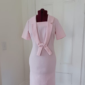 May include: A pink dress with a bow at the waist and a collar. The dress is on a mannequin in a room with white walls and a wooden floor.