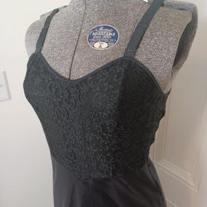 May include: A black slip dress with a lace bodice and adjustable straps. The dress is displayed on a gray dress form. The lace detailing adds a touch of elegance to the classic design. The dress is a size A.