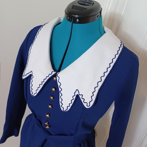 May include: A vintage navy blue dress with a white, scalloped collar and gold buttons. The dress features long sleeves and a matching belt. The collar is trimmed with navy blue stitching. The dress is displayed on a teal dress form.