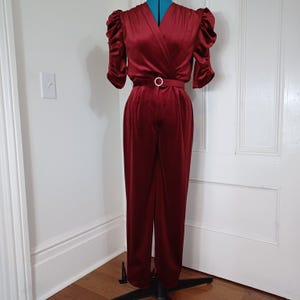 May include: A burgundy jumpsuit with a wrap-style bodice and short, puffed sleeves. The jumpsuit features a matching belt with a circular buckle. The fabric has a slight sheen, and the trousers are straight-legged.