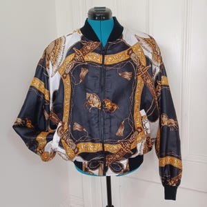 May include: A black, white, and gold bomber jacket with a detailed equestrian-themed pattern. The jacket features a zip-up front, black ribbed cuffs, and a black ribbed hem. The design includes horses, tassels, and decorative elements.