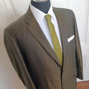 May include: A tailored olive green suit displayed on a mannequin. The suit includes a white dress shirt, a patterned green tie, and a white pocket square. The suit jacket has a two-button closure.