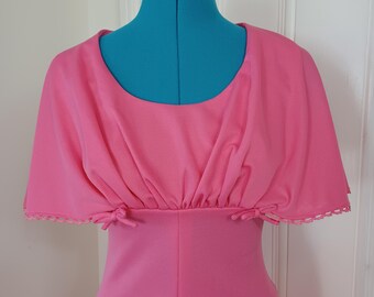 May include: A bright pink dress with a cape-like overlay. The dress features a scoop neckline and a fitted bodice. The cape has a scalloped edge and is gathered at the waist with small bows. The dress is displayed on a blue dress form.