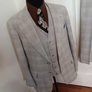 May include: A three-piece suit featuring a light gray blazer and matching vest with a subtle plaid pattern. The ensemble includes a dark brown button-down shirt and a dark brown necktie with a white and light blue intricate floral pattern.