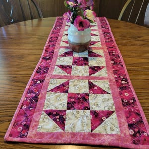 May include: A quilted table runner with a pink and white floral pattern. The runner features a central design of alternating squares and triangles in shades of pink and white, framed by a darker pink border. A vase of flowers sits on top.