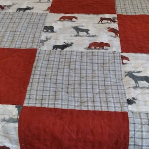 May include: A patchwork quilt featuring alternating red and patterned squares. The patterned squares display a white background with moose, bear, and deer illustrations. The quilt has a quilted diamond pattern.