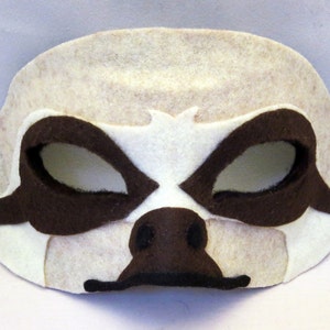 May include: A white felt sloth mask with brown felt eyes and a nose. The mask has a black elastic band for wearing.