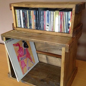 Peut inclure: Une étagère en bois à deux niveaux. Le niveau supérieur a une rangée de CD et le niveau inférieur a un album de disque avec le titre "Captain Beefheart" visible. L'étagère est en bois de récupération et a un aspect rustique.