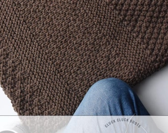 Puede incluir: Primer plano de una manta de punto marrón con un patrón texturizado. La manta está doblada, con las piernas de una persona en vaqueros visibles. El texto "KNITTING PATTERN" está en la parte inferior.