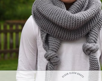 May include: A gray knitted scarf with a knotted fringe. The scarf is worn by a person and is draped around their neck. The text "Cluck Cluck Boots Knitting Pattern" is visible at the bottom of the image.