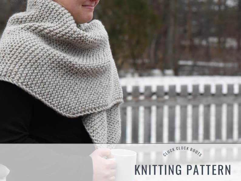 May include: A gray knitted shawl with a simple stitch pattern. The shawl is draped around a person's shoulders and is being held in place by their hand. The background is blurred and shows a wooden fence and snow.