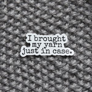 May include: A white sticker with the text "I brought my yarn just in case" sits on a gray knitted fabric background. The sticker's text is in a simple, sans-serif font. The knitted fabric has a textured, chunky appearance.