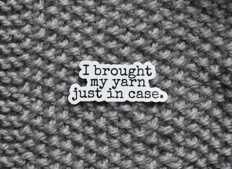 May include: A white sticker with the text "I brought my yarn just in case" sits on a gray knitted fabric background. The sticker's text is in a simple, sans-serif font. The knitted fabric has a textured, chunky appearance.