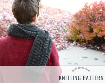 May include: A person wearing a dark gray knitted scarf with a textured pattern. The scarf is worn around the neck and draped over the shoulder. The person is standing in front of a background of fall foliage.