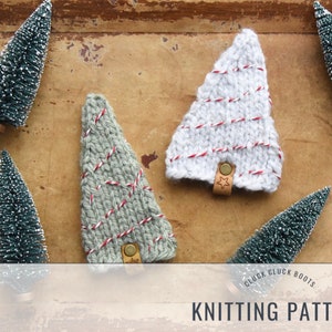 May include: Two knitted Christmas tree ornaments, one white with red stripes and one green with red stripes. Both ornaments have a small star charm attached. The ornaments are on a wooden surface with small artificial Christmas trees.