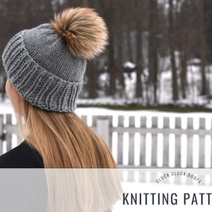 Puede incluir: Gorro de punto gris con un pompón marrón esponjoso. El gorro tiene un patrón de canalé y lo lleva una persona con el pelo largo y rubio. El fondo es una escena de invierno nevada con una valla blanca de madera.