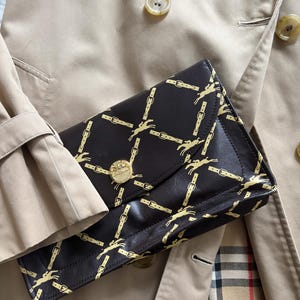 May include: A dark brown clutch bag featuring a gold equestrian design and a gold button clasp. The bag is placed on a beige trench coat.