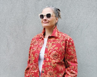 Könnte beinhalten: Rote Jacke mit floralem Muster und Knopfleiste. Die Jacke hat ein Muster aus großen, goldbraunen Blumen und grünen Blättern. Die Person auf dem Bild trägt eine Sonnenbrille und ein weißes Oberteil unter der Jacke.