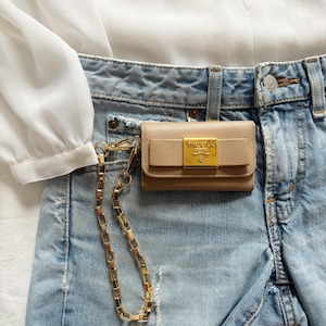 Peut inclure: Un portefeuille en cuir beige avec une chaîne dorée attachée, posé sur un short en jean bleu clair. Le portefeuille porte un logo doré "PRADA". Une blouse blanche est visible en arrière-plan.