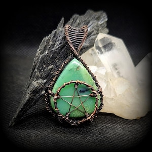 Puede incluir: Un colgante de piedra verde con un diseño de pentagrama envuelto en alambre de cobre. El colgante está sobre un fondo blanco y negro.