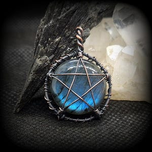 May include: A handcrafted pendant with a round, iridescent blue gemstone in a wire frame. A copper pentagram is overlaid on the stone, and the pendant is suspended from a twisted copper bail. The pendant is set against a dark background.