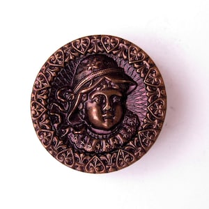 May include: A round, bronze-colored button with a detailed relief of a person wearing a hat and collar. The button's edge features an ornate, repeating pattern. The design is centered on a white background.
