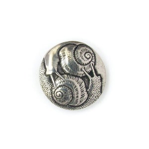 May include: A silver button with a raised design of two snails. The snails are facing each other and their shells are detailed with intricate swirls.
