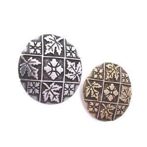 May include: Two round, decorative buttons. One is silver-toned, the other is gold-toned. Both feature a grid pattern with leaf and floral designs. The silver button has a slightly distressed appearance, while the gold button has a smooth finish.