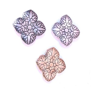 May include: Three decorative metal charms in the shape of a four-pointed star with intricate floral designs. The charms are in silver, copper, and rose gold.
