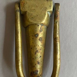May include: A brass door knocker in the shape of a vase with a decorative top and a ring handle. The knocker has a worn, aged patina.
