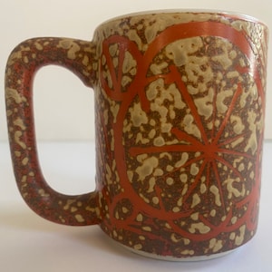 May include: A brown and tan ceramic mug with a red and white geometric design. The mug has a large handle.