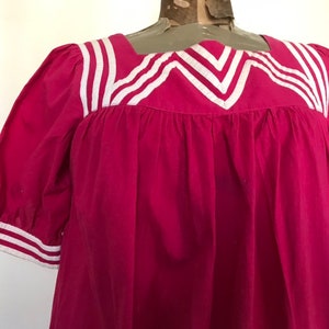 May include: A pink cotton dress with white trim around the neckline and sleeves. The neckline has a square shape with a V-shaped detail in the center.