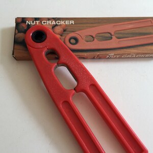 May include: A red plastic nutcracker with a handle and two cracking arms. The nutcracker is in its original packaging, which is brown with the words "NUT CRACKER" printed on it.