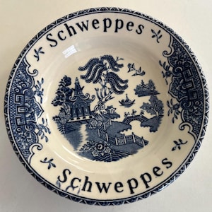 May include: A white ceramic saucer with a blue and white willow pattern and the word "Schweppes" written twice around the rim.