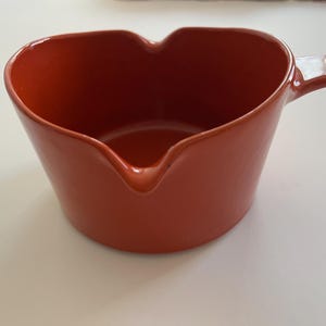 May include: A glossy, burnt orange ceramic saucepan shaped like a heart. The vessel features a smooth, solid-colored finish, a deep interior basin, a small pouring spout at the front, and a single handle extending from the right side of the rim.