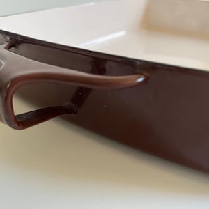 May include: Close-up of a rectangular brown baking dish with a white interior. The dish has curved handles on each side. The exterior is a glossy, dark brown color.