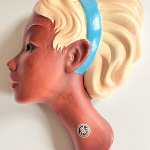 May include: A ceramic wall hanging in the shape of a woman's profile. The face is a reddish-brown color with blonde hair and a blue headband. The piece has a vintage aesthetic and a sticker with the letters "AK" on the neck.