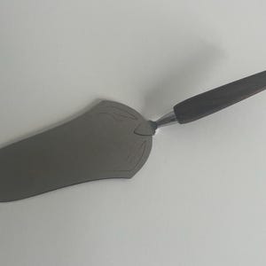 May include: A stainless steel cake server with a dark wooden handle. The server has a decorative design on the blade. The handle is angled for easy use. A kitchen utensil for serving cakes and pies.