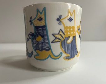 May include: White ceramic mug with a blue and yellow design featuring stylized animal figures. The animals are a bird, a cat, and a dog.
