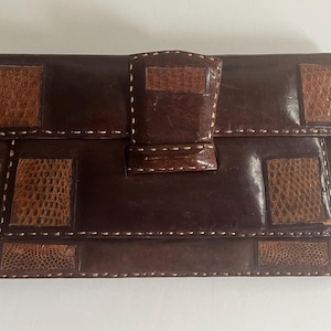 May include: A brown leather clutch purse with a geometric pattern of stitched leather squares. The purse has a flap closure with a stitched leather tab.