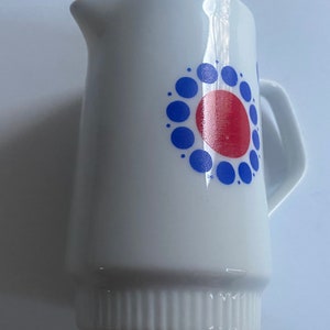 May include: White ceramic creamer with a blue and red geometric design. The design features a large red circle surrounded by a ring of smaller blue circles.