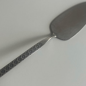 May include: A stainless steel cake server with a wide, oval blade and a long handle. The handle is decorated with a repeating pattern of small, stylized flower shapes. The server is designed for serving cakes and pastries.