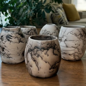Puede incluir: Colección de cinco jarrones de cerámica hechos a mano con un patrón único de rayos negros sobre fondo blanco. Los jarrones varían en forma y tamaño, con un acabado liso y brillante. Están dispuestos sobre una superficie de madera.